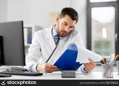 healthcare, medicine and people concept - male doctor with clipboard calling on desk phone at hospital. male doctor calling on desk phone at hospital