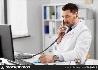healthcare, medicine and people concept - male doctor with clipboard calling on desk phone at hospital. male doctor calling on desk phone at hospital