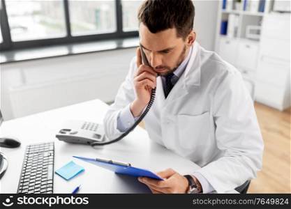 healthcare, medicine and people concept - male doctor with clipboard calling on desk phone at hospital. male doctor calling on desk phone at hospital