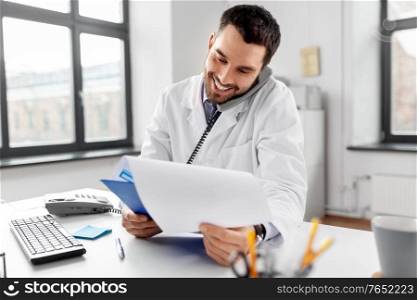 healthcare, medicine and people concept - smiling male doctor with clipboard calling on desk phone at hospital. male doctor calling on desk phone at hospital