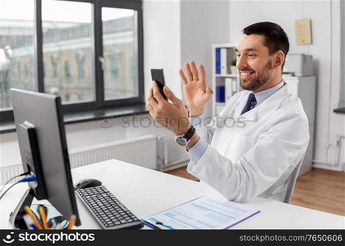healthcare, medicine and people concept - smiling male doctor with smartphone having video call and waving hand at hospital. doctor with smartphone having video call at clinic