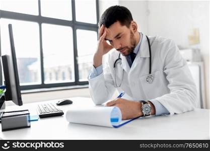 healthcare, medicine and people concept - stressed male doctor with clipboard at hospital. stressed male doctor with clipboard at hospital