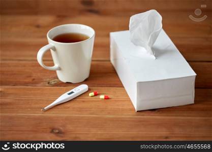 healthcare, medicine, flu and treatment concept - cup of tea, paper wipes and thermometer with pills