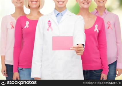 healthcare, people and medicine concept - close up of smiling women in shirts with pink breast cancer awareness ribbons and blank paper card over green background