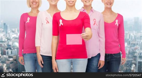 healthcare, people and medicine concept - close up of smiling women in shirts with pink breast cancer awareness ribbons and blank paper card over city background