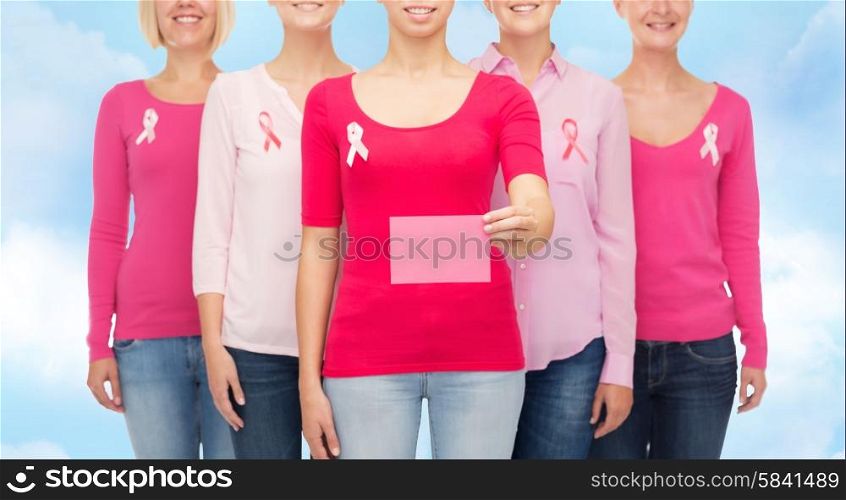 healthcare, people and medicine concept - close up of smiling women in shirts with pink breast cancer awareness ribbons and blank paper card over blue sky background