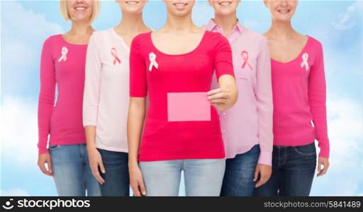 healthcare, people and medicine concept - close up of smiling women in shirts with pink breast cancer awareness ribbons and blank paper card over blue sky background