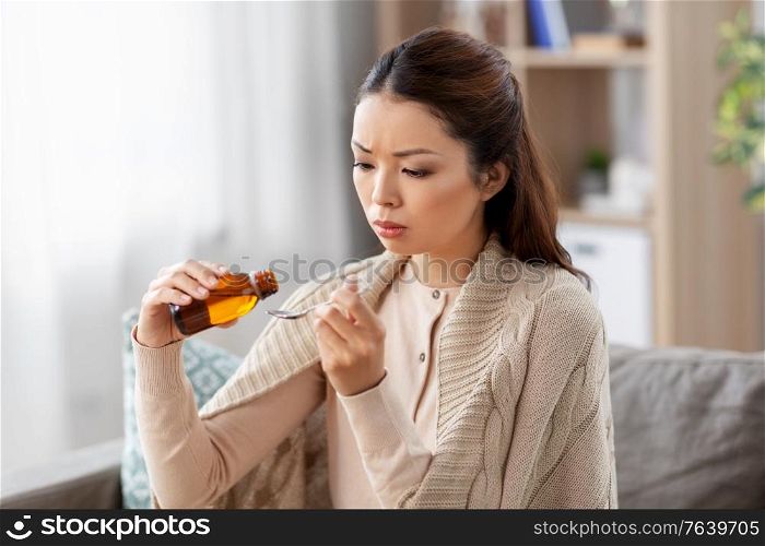 healthcare, people and medicine concept - sick asian woman pouring antipyretic or cough syrup from bottle to spoon at home. sick asian woman taking medicine at home
