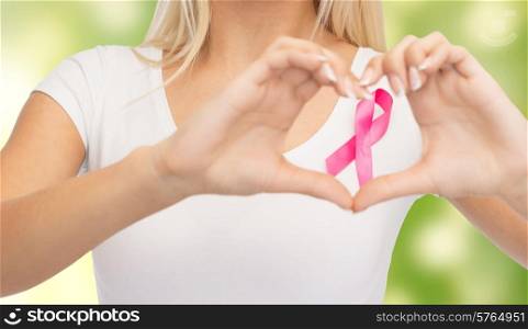 healthcare, people, charity and medicine concept - close up of young woman in blank white t-shirt with pink breast cancer awareness ribbon showing heart shape over green background