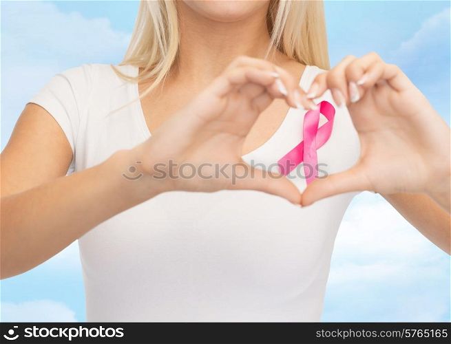 healthcare, people, charity and medicine concept - close up of young woman in blank white t-shirt with pink breast cancer awareness ribbon showing heart shape over blue sky background