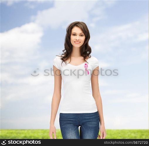 healthcare, people, charity and medicine concept - smiling young woman in t-shirt with pink breast cancer awareness ribbon over blue sky and grass background