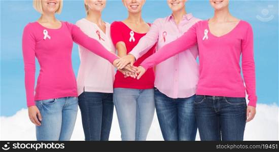 healthcare, people, gesture and medicine concept - close up of smiling women in blank shirts with pink breast cancer awareness ribbons putting hands on top over blue sky and white cloud background