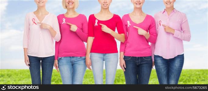 healthcare, people, gesture and medicine concept - close up of smiling women in blank shirts pointing fingers to pink breast cancer awareness ribbons over blue sky and grass background