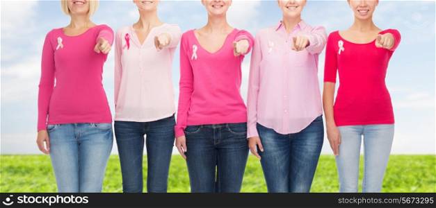 healthcare, people, gesture and medicine concept - close up of smiling women in blank shirts with pink breast cancer awareness ribbons pointing on you over blue sky and grass background