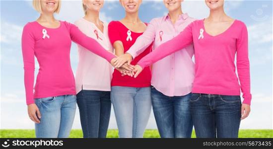 healthcare, people, gesture and medicine concept - close up of smiling women in blank shirts with pink breast cancer awareness ribbons putting hands on top over blue sky and grass background