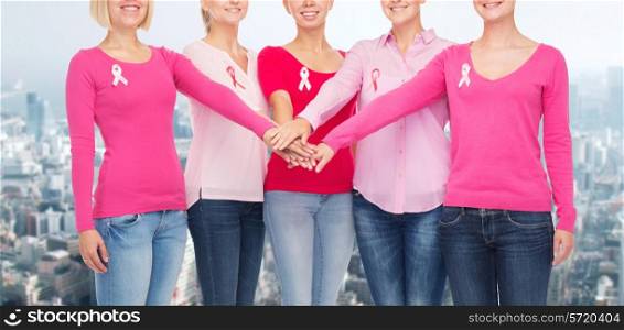 healthcare, people, gesture and medicine concept - close up of smiling women in blank shirts with pink breast cancer awareness ribbons putting hands on top over city background
