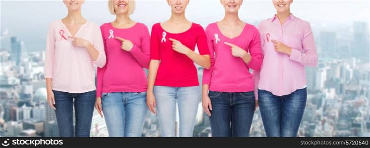 healthcare, people, gesture and medicine concept - close up of smiling women in blank shirts pointing fingers to pink breast cancer awareness ribbons over city background