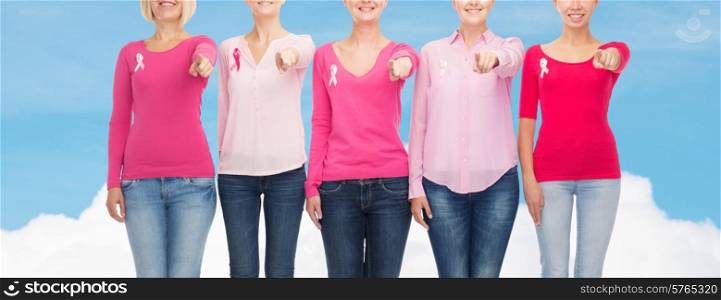 healthcare, people, gesture and medicine concept - close up of smiling women in blank shirts with pink breast cancer awareness ribbons pointing on you over blue sky and white cloud background