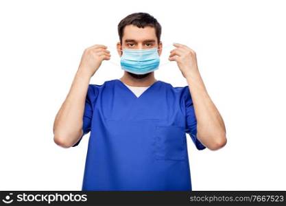 healthcare, profession and medicine concept - doctor or male nurse in blue uniform putting on face protective medical mask for protection from virus disease over white background. male doctor in blue uniform putting on mask