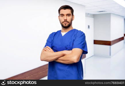 healthcare, profession and medicine concept - doctor or male nurse in blue uniform with crossed arms over hospital corridor background. doctor or male nurse in blue uniform