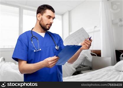 healthcare, profession and medicine concept - doctor or male nurse in blue uniform with cardiogram on clipboard over hospital ward on background. male doctor with cardiogram on clipboard