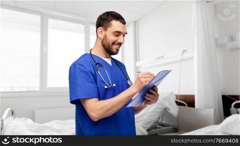 healthcare, profession and medicine concept - happy smiling doctor or male nurse in blue uniform writing medical report on clipboard over hospital ward on background. male doctor writing medical report on clipboard