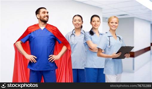 healthcare, profession and medicine concept - happy smiling male doctor in blue uniform and red superhero cape and female nurses over hospital corridor background. male doctor in superhero cape and nurses at clinic