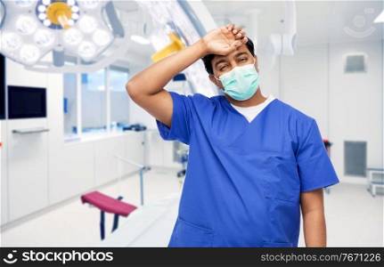 healthcare, profession and medicine concept - tired indian doctor or male nurse in blue uniform and face medical mask for protection from virus disease over surgery at hospital on background. tired indian male doctor in blue uniform and mask