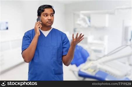 healthcare, stomatology and medicine concept - indian doctor or male dentist in blue uniform with stethoscope calling on smartphone over dental clinic office background. indian doctor calling on phone at dental office