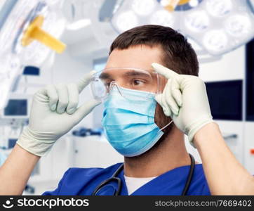 healthcare, surgery and medicine concept - doctor or male nurse in blue uniform, face protective medical mask for protection from virus disease, goggles and gloves over hospital on background. male doctor in goggles, mask and gloves