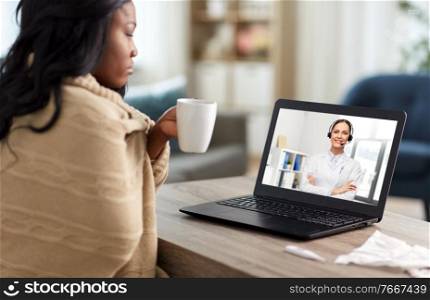 healthcare, technology and people concept - african american sick woman in blanket having video call with doctor on laptop computer and drinking tea at home. sick woman having video call with doctor on laptop
