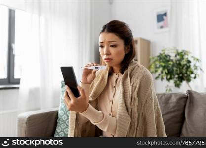 healthcare, technology and people concept - sick asian woman in blanket with thermometer having video call on smartphone at home. sick woman having video call on smartphone
