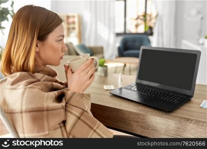 healthcare, technology and people concept - sick young woman in blanket drinking tea and having video call on laptop computer at home. sick woman with tea having video call on laptop