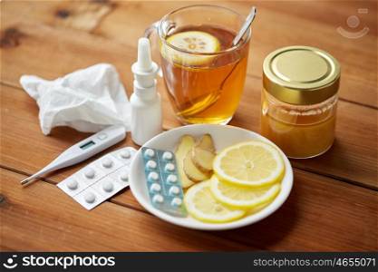 healthcare, traditional medicine and flu concept - tea cup with lemon, ginger, honey and pills on wooden table