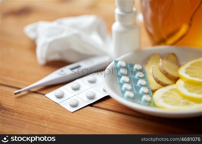 healthcare, traditional medicine and flu concept - tea cup with lemon, ginger, honey and pills on wooden table