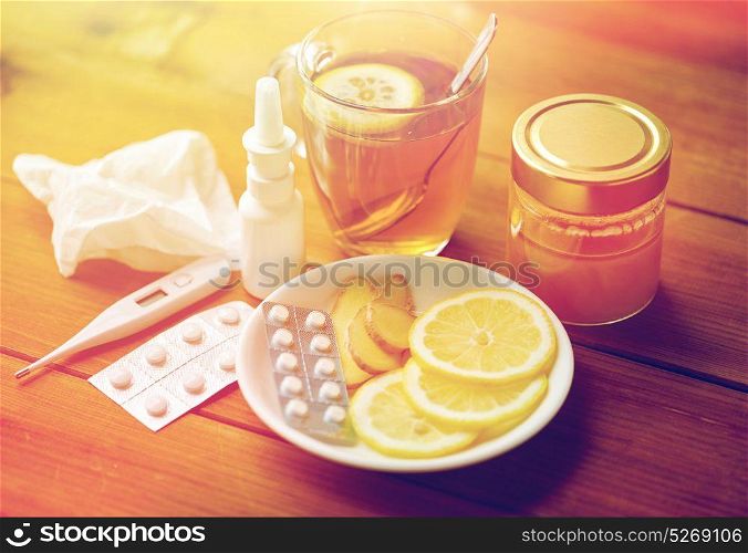 healthcare, traditional medicine and flu concept - tea cup with lemon, ginger, honey and pills on wooden table. traditional medicine and drugs