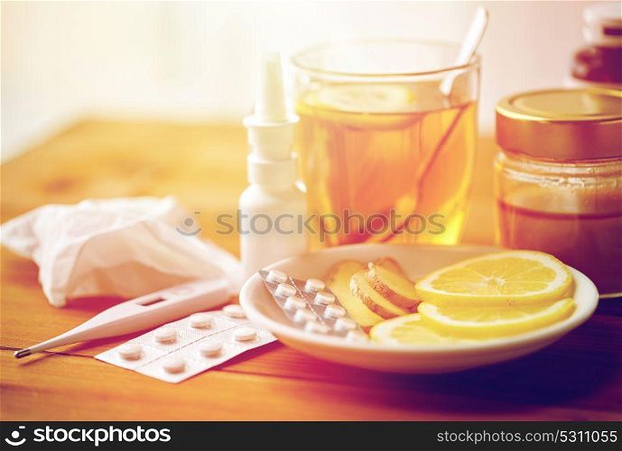 healthcare, traditional medicine and flu concept - tea cup with lemon, ginger, honey and pills on wooden table. traditional medicine and drugs