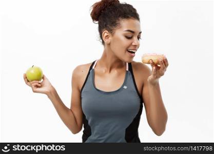 Healthy and diet concept - Beautiful sporty African American make a decision between donut and green apple. Isolated on white background. Healthy and diet concept - Beautiful sporty African American make a decision between donut and green apple. Isolated on white background.