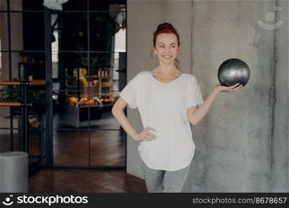 Healthy and happy young ginger woman holds small silver fitball on open palm of one hand against background of gray wall and glass door in fitness studio, smiling at camera. Sport and pilates concept. Red haired happy woman standing in gym with small silver ball