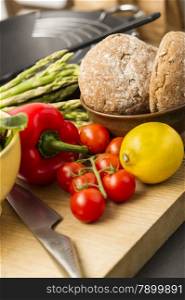 Healthy assortment of fresh vegetables including red bell pepper, lemon, asparagus tips and tomatoes with wholegrain rolls on a wooden kitchen counter with a sharp knife