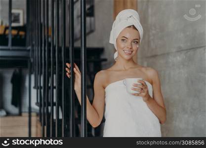 Healthy attractive European woman with pensive expression smiles gently, applies face cream on complexion, stands wrapped in towel, holds mug of drink, relaxes at home. Natural beauty concept