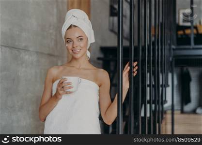 Healthy attractive European woman with pensive expression smiles gently, applies face cream on complexion, stands wrapped in towel, holds mug of drink, relaxes at home. Natural beauty concept