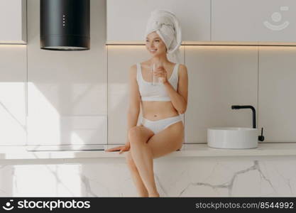 Healthy beauty. Slim sportive young girl sitting on white marble kitchen table top in white underwear while drinking glass of mineral pure water in morning, enjoying new day. Water balance concept. Slim sportive young girl sitting on white marble kitchen table top and drinking glass of mineral pure water