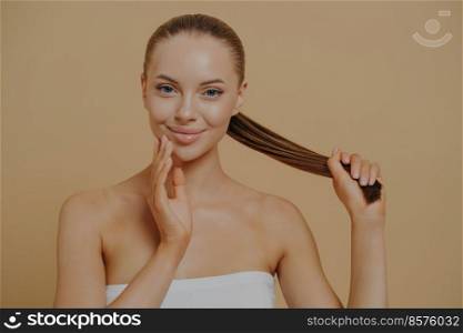 Healthy beauty. Young attractive lady with naked shoulders on beige background, enjoying her soft clean skin, touching face and holding strong damage-free hair in ponytail. Haircare and skincare. Beauty portrait of young naturally beautiful woman touching facial skin and holding hair in ponytail