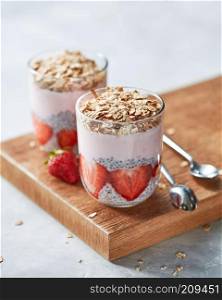 Healthy breakfast from natural ingredients - homemade granola, halves of strawberries, chia seeds and yogurt in a glasses with a spoons on a wooden board on a gray concrete background, copy space.. Homemade oat granola with strawberries and in glasses with slices of strawberries, chia seeds on wooden background. Copy space.