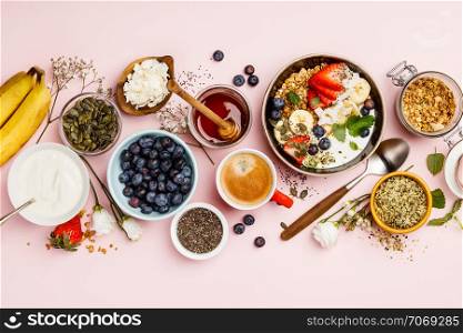 Healthy breakfast set. Oat granola with fresh berries, banana, yogurt, maple syrup, seeds and mint leaves with cup of coffee on pink background, flat lay, top view