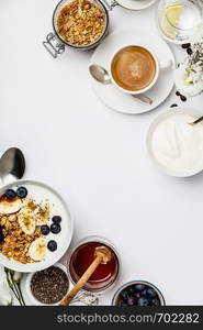 Healthy breakfast set. Oat granola with yogurt, honey, fresh bananas, blueberries, chia seeds in bowl and cup of coffee on white background, top view, copy space. Healthy breakfast set on white background, top view, copy space