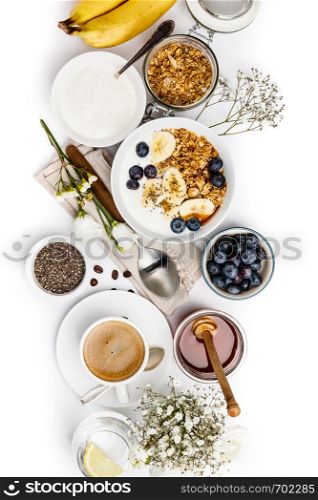 Healthy breakfast set. Oat granola with yogurt, honey, fresh bananas, blueberries, chia seeds in bowl and cup of coffee on white background, top view. Healthy breakfast set on white background, top view, copy space