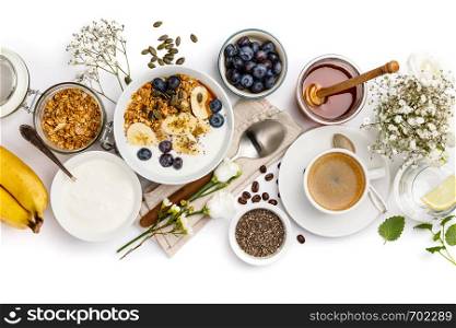 Healthy breakfast set. Oat granola with yogurt, honey, fresh bananas, blueberries, chia seeds in bowl and cup of coffee on white background, top view. Healthy breakfast set on white background, top view, copy space