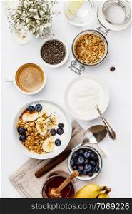 Healthy breakfast set. Oat granola with yogurt, honey, fresh bananas, blueberries, chia seeds in bowl and cup of coffee on white background, top view